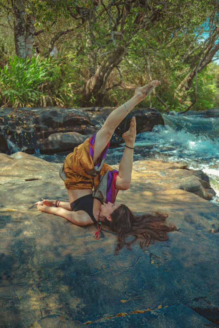 Woman performing yoga on a rock by a flowing river amidst a lush forest setting.